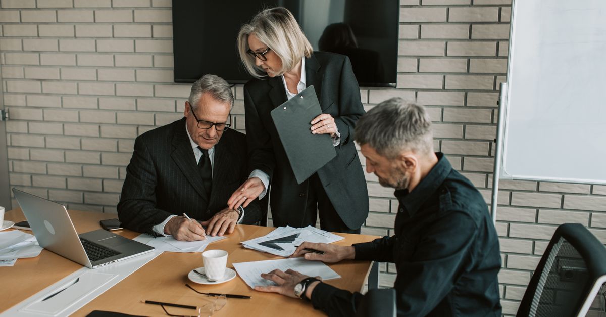 Three mature professionals in a business meeting discussing and signing documents in an office setting.