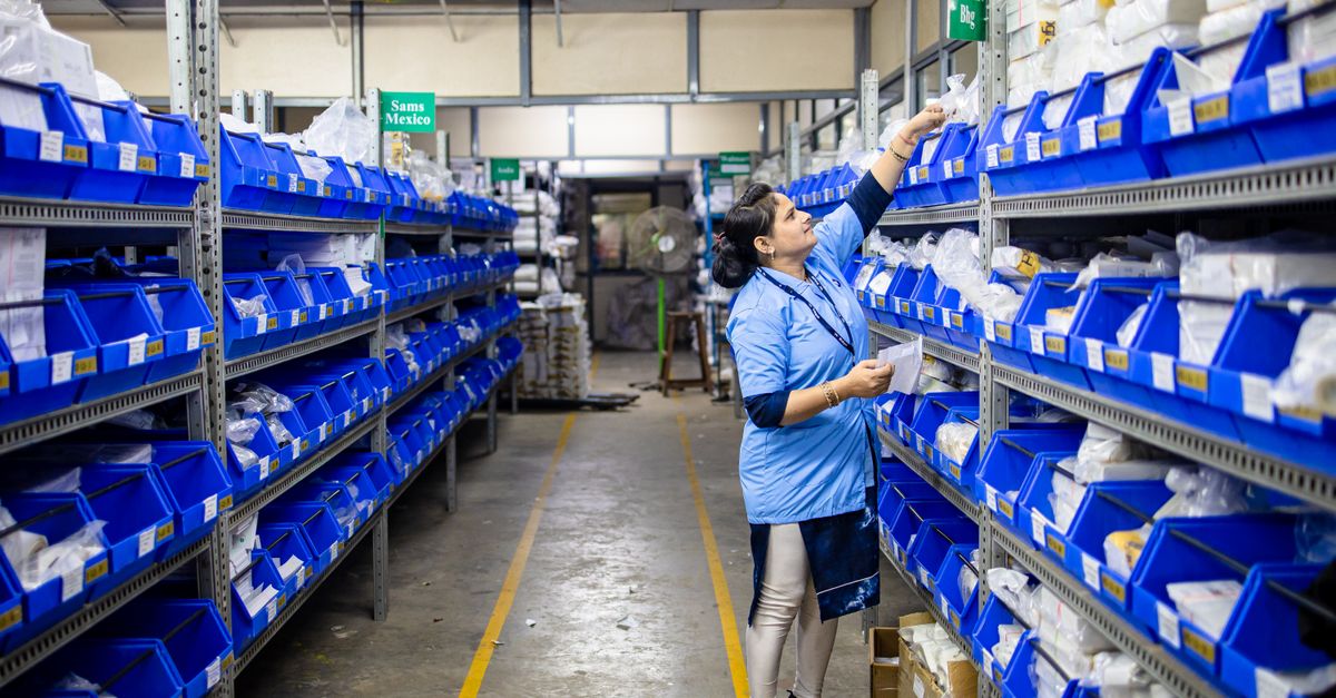 Woman in uniform organizing inventory in a warehouse aisle with labeled bins.