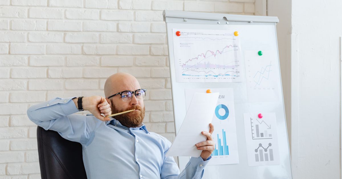 A businessman in glasses reviews financial charts, deep in thought by a whiteboard with graphs.