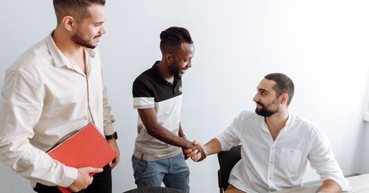 Three men in an office setting, shaking hands, representing teamwork and diversity.