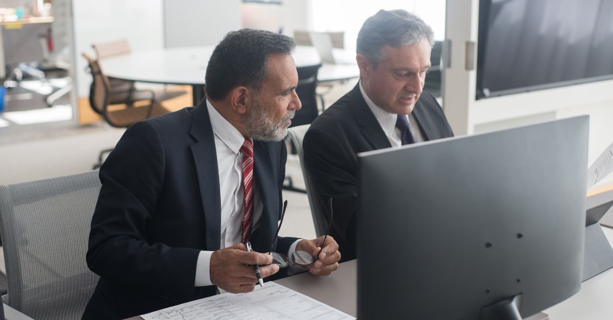 Two male executives in office discussing documents at a computer workstation.